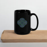 On top of a cutting board, a Xigfireon black ceramic 15oz mug featuring the Living Colour iteration of the `Reach Of The Spirit` Celtic knot design. This Celtic knot symbolizes Mother Earth.