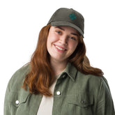 A young woman wearing an olive green Xigfireon vintage corduroy cap embroidered with the Living Colour iteration of the `Reach Of The Spirit` Celtic knot design.
