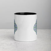 A tabletop displaying a Xigfireon white ceramic 11oz mug featuring the Living Colour iteration of the `Reach Of The Spirit` Celtic knot design, with black handle and inside. This Celtic knot symbolizes Mother Earth.