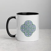 A table top displaying a Xigfireon white ceramic 11oz mug featuring the Living Colour iteration of the `Reach Of The Spirit` Celtic knot design, with black handle and inside. This Celtic knot symbolizes Mother Earth.