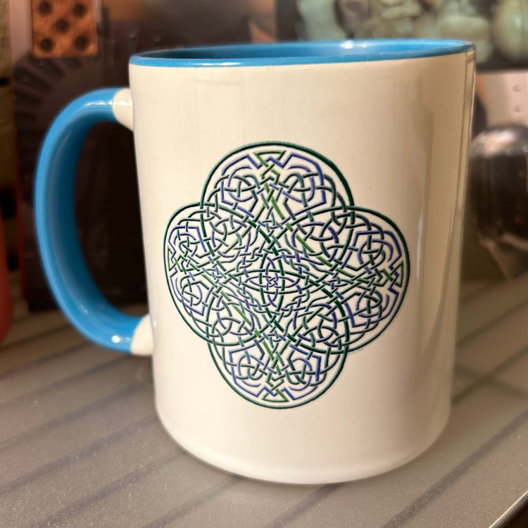 A Xigfireon white ceramic 11oz mug featuring the Living Colour iteration of the `Reach Of The Spirit` Celtic knot design, with blue handle and inside. This Celtic knot symbolizes Mother Earth.