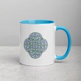 A tabletop displaying a Xigfireon white ceramic 11oz mug featuring the Living Colour iteration of the `Reach Of The Spirit` Celtic knot design, with blue handle and inside. This Celtic knot symbolizes Mother Earth.