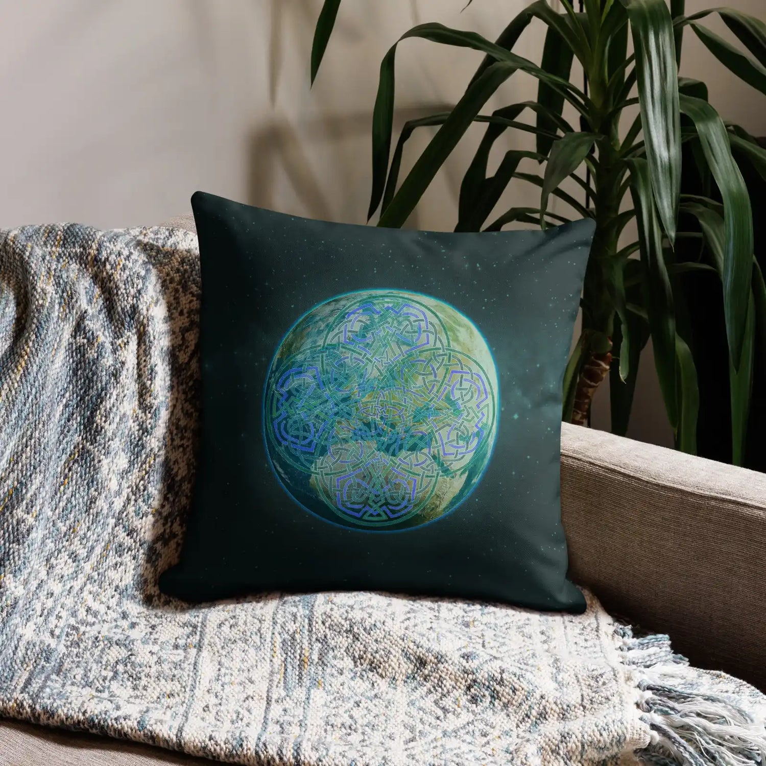 An 18 inch by 18 inch Xigfireon pillow case featuring the Sacred Planet iteration of the `Reach Of The Spirit` Celtic knot design, symbolizing Mother Earth. It is shown placed at the end of a sofa besides a tall house plant.