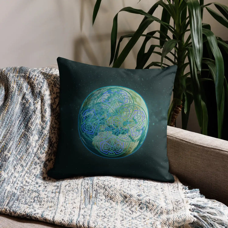 An 18 inch by 18 inch Xigfireon pillow case featuring the Sacred Planet iteration of the `Reach Of The Spirit` Celtic knot design, symbolizing Mother Earth. It is shown placed at the end of a sofa besides a tall house plant.