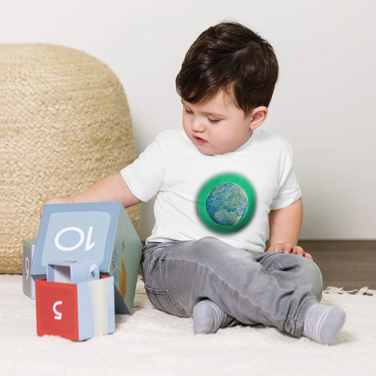 A young boy shown wearing a white Xigfireon toddler graphic t-shirt featuring the Deep Forest iteration of the `Reach Of The Spirit` Celtic knot design. The `Reach Of The Spirit` Celtic knot symbolizes Mother Earth.