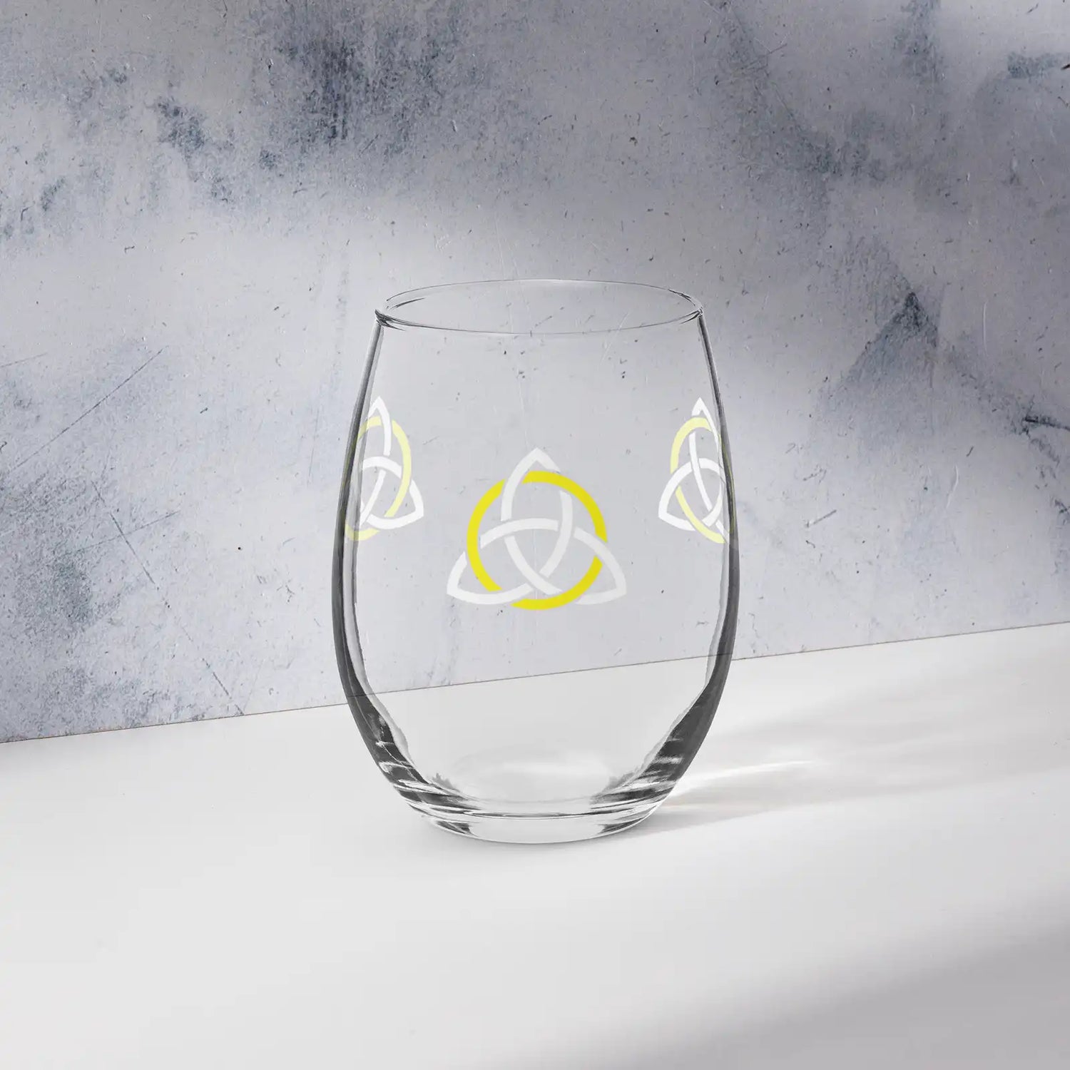 The front side of a stemless wine glass featuring the White & Yellow iteration of the Trinity Knot design shown on a counter in front of a textured wall. Also referred to as the triquetra, the Trinity Knot has long symbolized unity and balance.