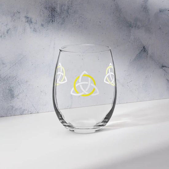 The front side of a stemless wine glass featuring the White & Yellow iteration of the Trinity Knot design shown on a counter in front of a textured wall. Also referred to as the triquetra, the Trinity Knot has long symbolized unity and balance.