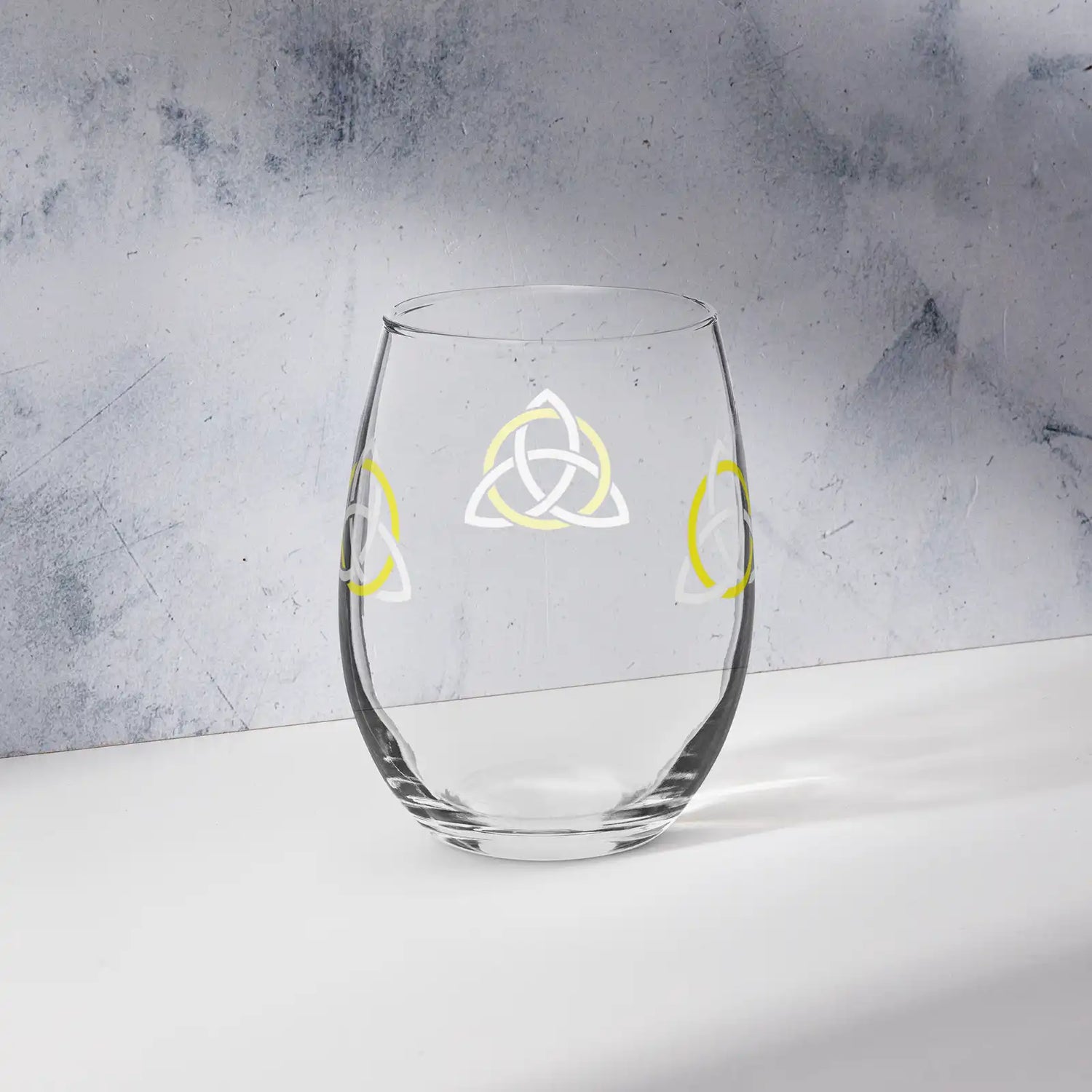 The back side of a stemless wine glass featuring the White & Yellow iteration of the Trinity Knot design shown on a counter in front of a textured wall. Known as the triquetra, the Trinity Knot embodies unity and balance.