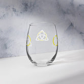 The back side of a stemless wine glass featuring the White & Yellow iteration of the Trinity Knot design shown on a counter in front of a textured wall. Known as the triquetra, the Trinity Knot embodies unity and balance.