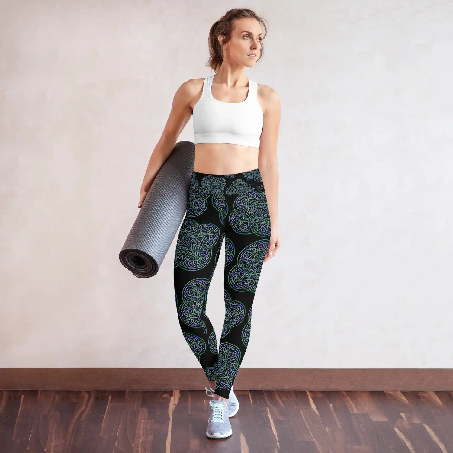 A young woman holding a yoga mat and wearing a pair of Xigfireon graphic yoga leggings featuring the Living Colour Black patterned series of the Reach Of The Spirit Celtic knot design.
