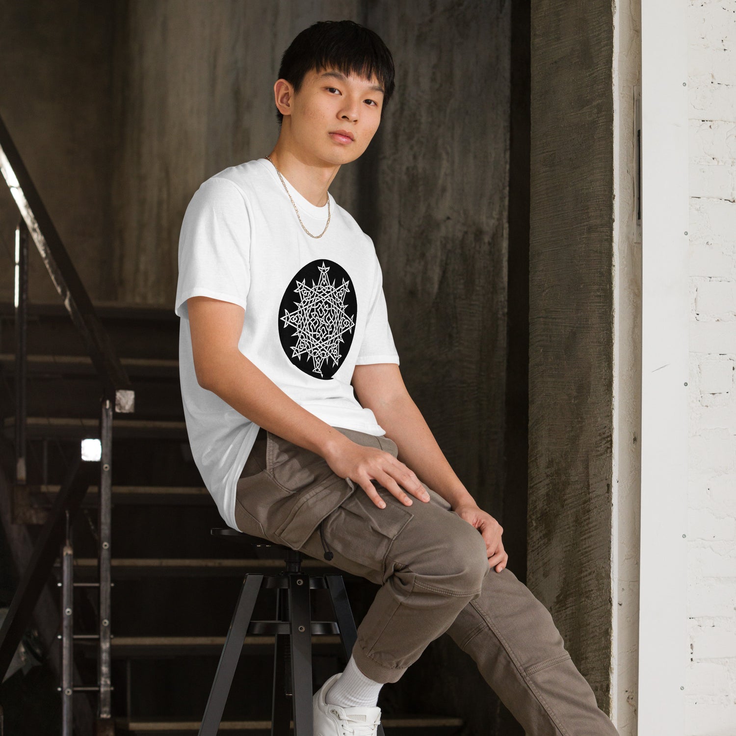 A young man, sitting on a stool, wearing a white Xigfireon graphic t-shirt featuring the Black & White Circle iteration of the `Morning Star Fire` Celtic knot design. The `Morning Star Fire` Celtic knot represents Father Sun and the eternal Cosmic Light.