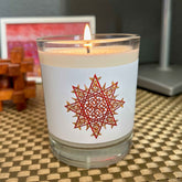 A nightstand displaying a Xigfireon glass jar soy wax candle featuring the Fire Colour iteration of the `Morning Star Fire` Celtic knot design. The `Morning Star Fire` Celtic knot symbolizes Father Sun.