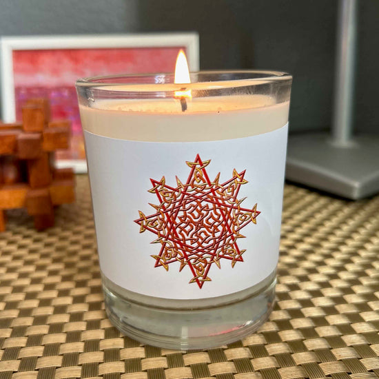 A nightstand displaying a Xigfireon glass jar soy wax candle featuring the Fire Colour iteration of the `Morning Star Fire` Celtic knot design. The `Morning Star Fire` Celtic knot symbolizes Father Sun.