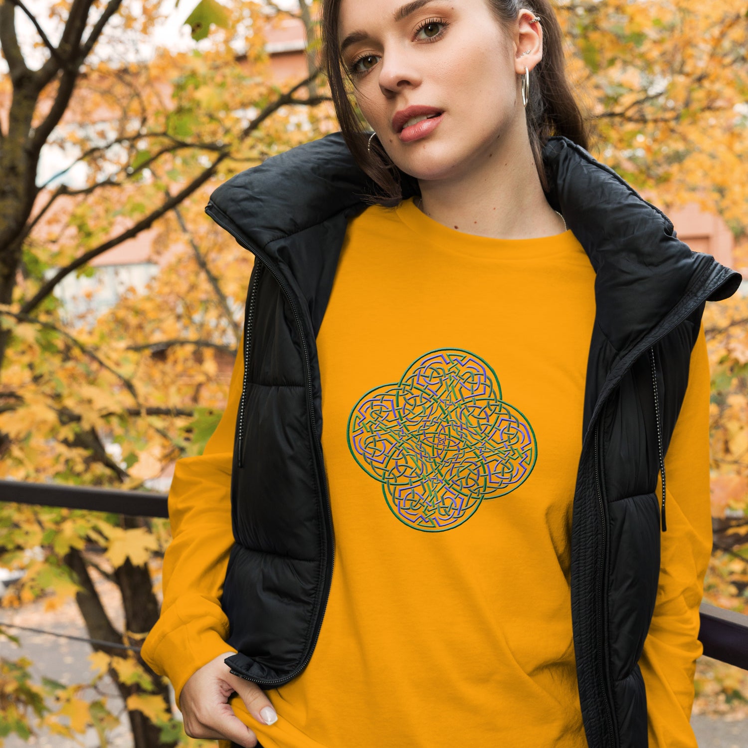 A young woman wearing a gold Xigfireon long sleeve graphic t-shirt featuring the Living Colour iteration of the `Reach Of The Spirit` Celtic knot design. The `Reach Of The Spirit` Celtic knot symbolizes Mother Earth.